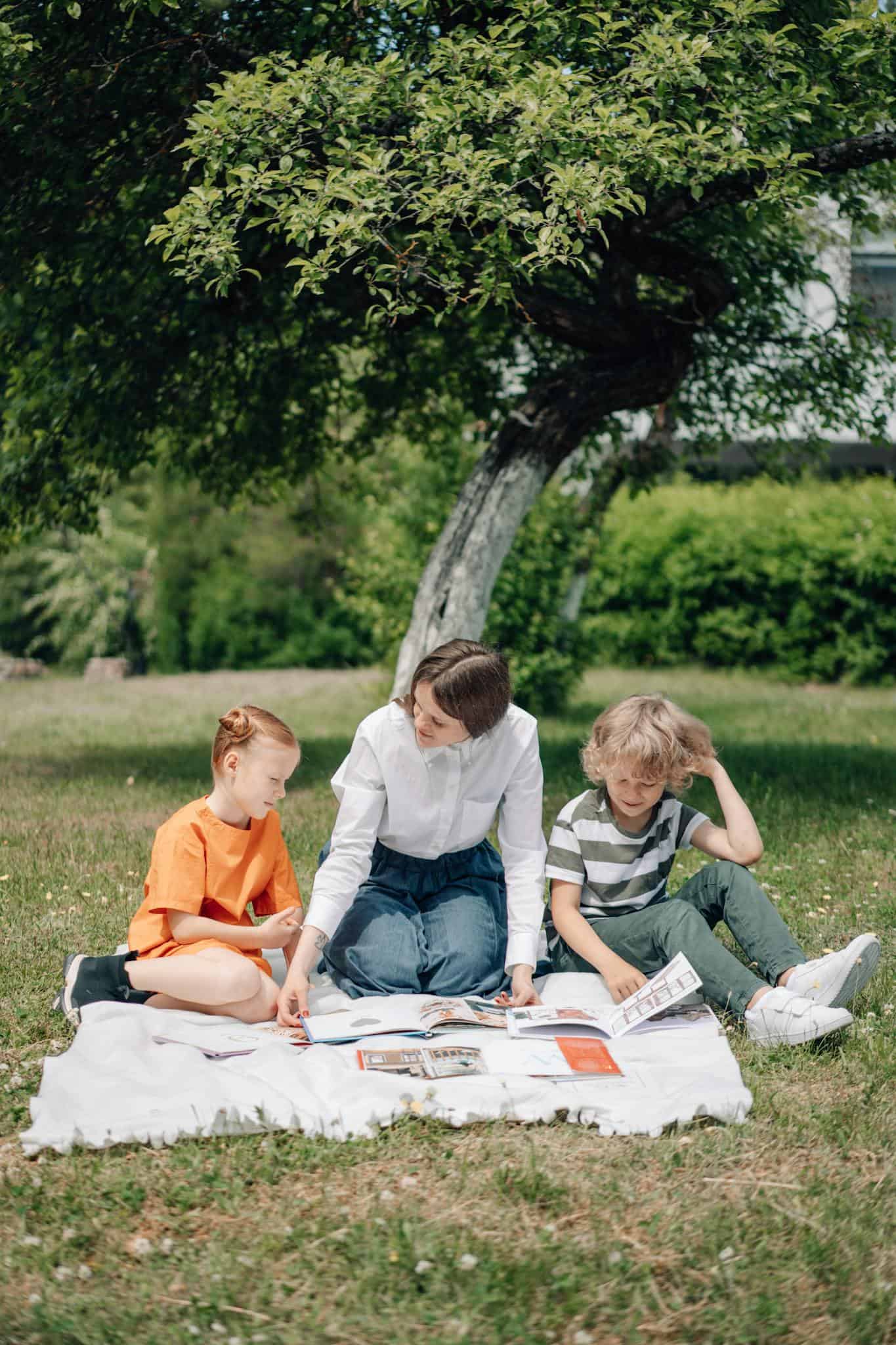 A woman teaches two kids outdoors on a picnic blanket under a tree.